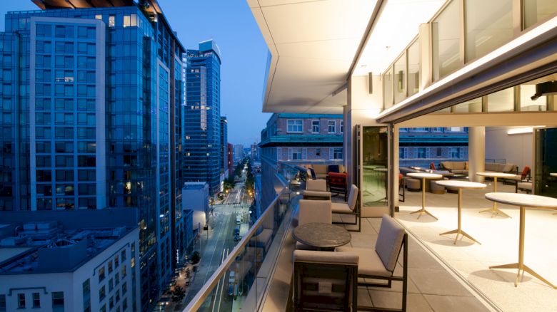 A modern balcony overlooks a city street with tall buildings, featuring outdoor seating under soft lighting in an urban setting.