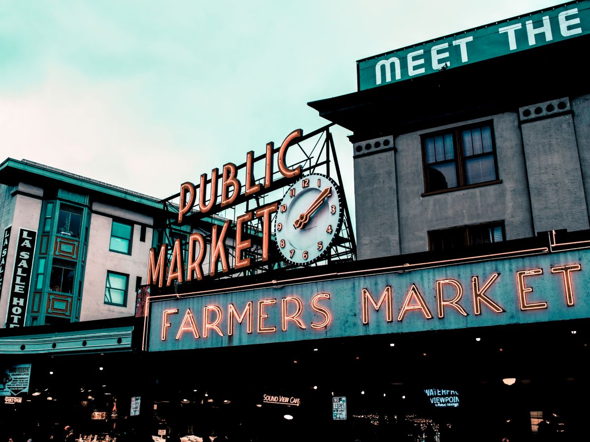 A public market sign with a clock and "Farmers Market" in neon lights on a building with "MEET THE" partially visible above.