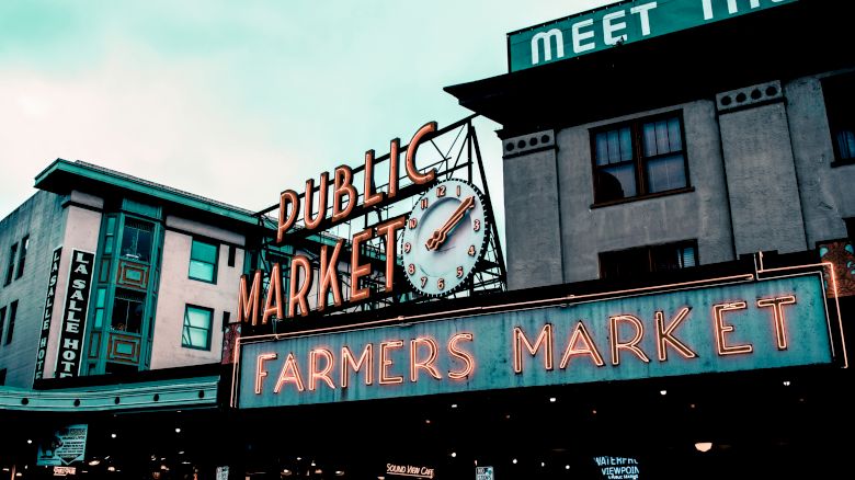 A public market sign with a clock and "Farmers Market" in neon lights on a building with "MEET THE" partially visible above.