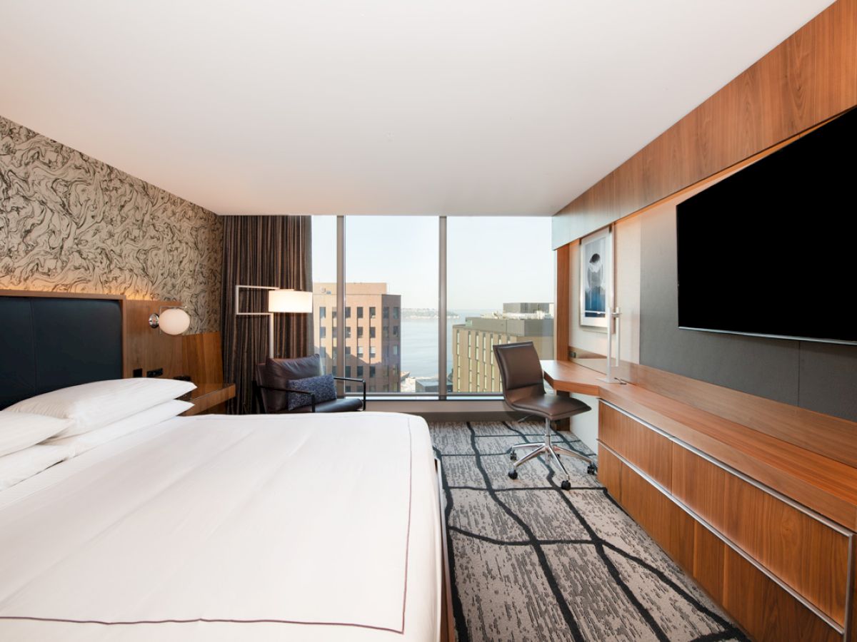 The image shows a modern hotel room with a large bed, desk, chair, and TV, featuring a city view through a floor-to-ceiling window.