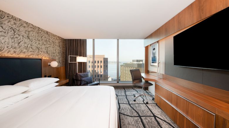 The image shows a modern hotel room with a large bed, desk, chair, and TV, featuring a city view through a floor-to-ceiling window.