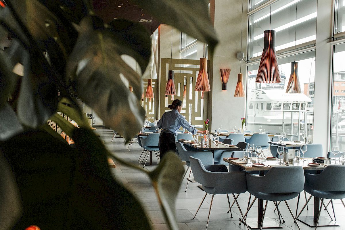 A modern restaurant with blue chairs and copper pendant lights, featuring a person setting a table under natural light.