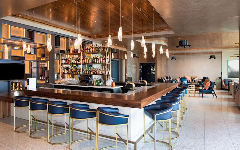 A modern bar with stylish lighting, a well-stocked counter, and blue barstools. Cozy seating area visible in the background.