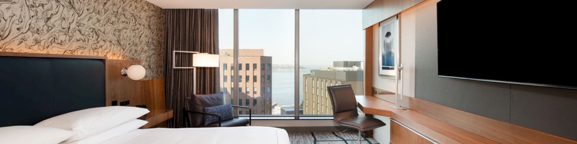 A modern hotel room with a large bed, flat-screen TV, desk, and a window view of a cityscape.